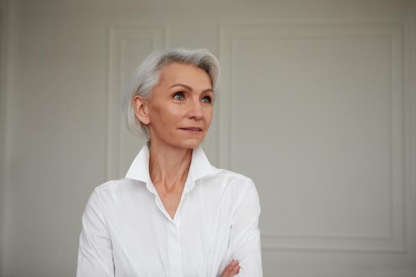 Portrait of an adult elderly self-confident woman with white hair looking awey into distance in white shirt hands crossed background wall