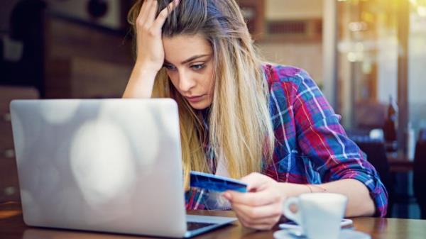 A young woman sitting at her laptop while holding her credit card with a stressed look on her face